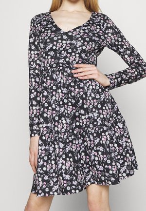 Frau trägt ein schwarzes, langärmeliges Kleid mit pinkem, weißem und violettem Blumenmuster und posiert mit einer Hand auf der Hüfte.