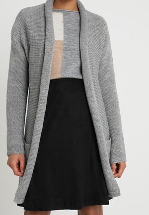 Žena v dlouhém šedém pleteném kardiganu přes šedé tričko s barevnými bloky a černou sukni střihu A-line.