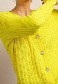 Cardigan a maglia giallo lime con texture a coste, scollatura a V e bottoni floreali scintillanti sul davanti, che mostra una vestibilità aderente.