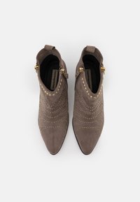 Dos botas de tobillo grises con punta afilada, adornadas con tachuelas doradas y cremalleras a los lados, sobre un fondo blanco liso.