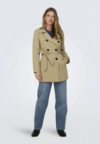 Beige Trenchcoat mit doppelreihigen Knöpfen, tailliertem Gürtel und Seitentaschen; kombiniert mit einem blauen Oberteil und weit geschnittenen Jeans.