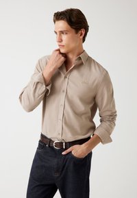 Beige overhemd met lange mouwen en knoopsluiting, met een subtiele textuur, voorzien van een klein logo en een standaard kraag, gecombineerd met donkerdenim jeans.
