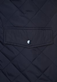 Marineblau gesteppte Jacke mit einer rechteckigen Tasche, die mit einem glänzenden schwarzen Druckknopf gesichert ist und eine elegante, glatte Stoffoberfläche zeigt.