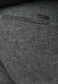 Grijze broeken met een houndstooth-patroon, voorzien van een achterzak met een zwarte leren label. De stof heeft een gestructureerde afwerking.