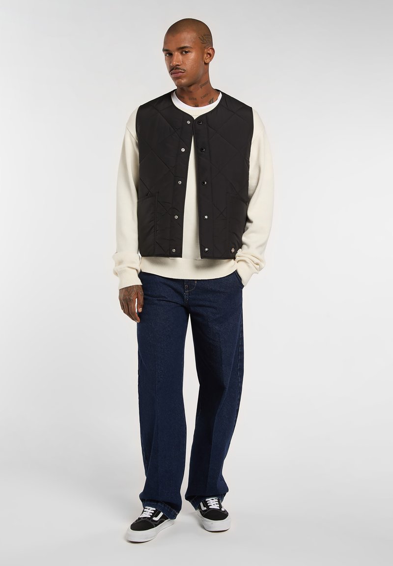 Sort quiltet vest med trykknapper, båret over en hvid sweater. Parret med løse mørkeblå jeans og sorte sneakers med hvide detaljer.