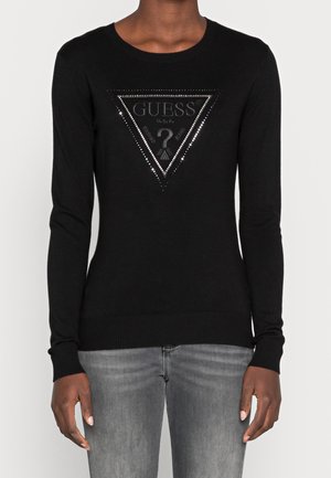Schwarzer figurbetonter Langarmpullover mit silbernen Nieten am dreieckigen Guess-Logo auf der Brust, kombiniert mit ausgewaschenen grauen Jeans.