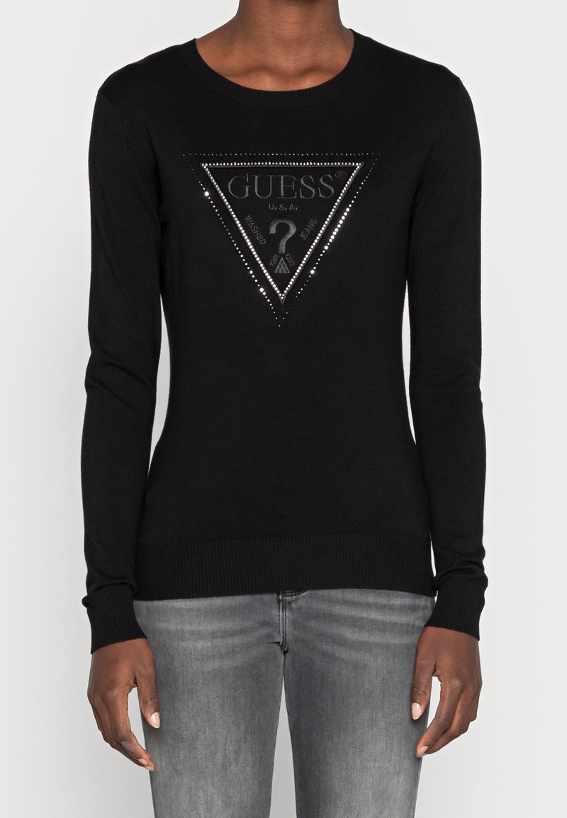 Pull noir ajusté à manches longues avec logo triangulaire Guess clouté d'argent sur la poitrine, porté avec un jean gris délavé.