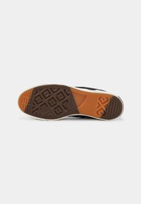 Semelle d'une basket noire et blanche montrant un profil en caoutchouc brun et orange avec des motifs géométriques et un embossage du logo sur fond blanc.