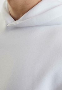 Felpa bianca in cotone con una texture morbida, dotata di cappuccio con cordino e scollatura con vestibilità rilassata. Il tessuto appare liscio e caldo.