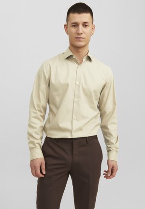 Chemise beige à manches longues avec col, fabriquée en tissu lisse ; associée à un pantalon marron ajusté, avec une coupe slim.
