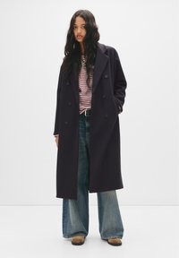 Cappotto doppiopetto di un blu navy scuro con ampi risvolti, abbinato a una camicia a righe e jeans a gamba larga blu. La texture appare liscia e sartoriale.