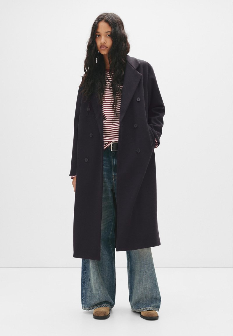 Cappotto doppiopetto di un blu navy scuro con ampi risvolti, abbinato a una camicia a righe e jeans a gamba larga blu. La texture appare liscia e sartoriale.