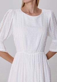 Robe blanche avec des manches bouffantes, tissu texturé présentant des motifs à pois, accents en dentelle le long du corps et des manches, et une jupe plissée.