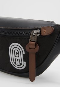 Černá nylonová pásková taška s hladkým koženým lemováním, vybavená stříbrným zipem a výraznou bílou nášivkou s logem. Hnědé akcenty na táhle zipu.