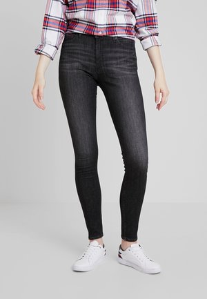 Persoon die zwarte skinny jeans, witte sneakers en een rood-wit-blauw geruite blouse met opgerolde mouwen draagt tegen een effen achtergrond.