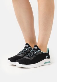 Legs wearing black and turquoise Skechers sneakers with white soles, standing on a white surface.