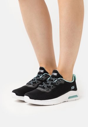 Des jambes portant des baskets noires et turquoise de Skechers avec des semelles blanches, debout sur une surface blanche.