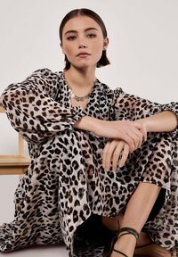 Durchscheinendes Tiermusterkleid mit langen Puffärmeln in Beige und Schwarz, ausgestattet mit einem tiefen Ausschnitt und fließender Silhouette. Das Model sitzt und trägt eine mehrlagige Halskette.