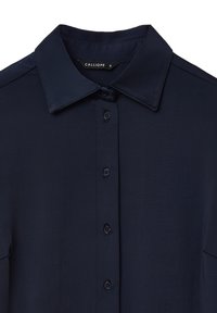 Camicia blu navy con colletto, chiusura anteriore con bottoni e maniche corte. Tessuto liscio e leggero luccichio. Etichetta del marchio visibile.