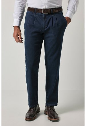 Mand iført mørkeblå plisserede bukser, brun læderbælte, hvid skjorte og mørkebrune brogues står med den ene hånd i lommen.