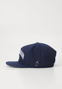 Mørkeblå baseballcap med flad skygge, der har "JORDAN" i hvid tekst foran og et gråt broderet Jumpman-logo på siden.