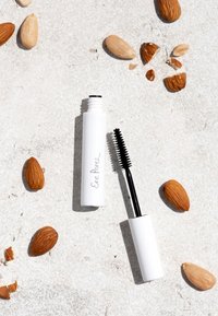 Tube de mascara blanc avec brosse noire, arborant un logo en script simple, placé parmi des amandes entières et cassées sur une surface texturée claire.