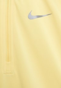 Vaaleankeltainen urheilupaita, jossa on puoli vetoketju. Vasemmalla puolella on heijastava hopeanvärinen Nike-logo ja kangas on sileä ja tekstuuri.