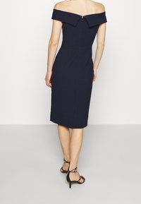 Námořnické šaty s odhalenými rameny, se tapered siluetou, zapínáním na zip, po kolena a s rozparkem na zadní straně. Nosené s černými sandály na podpatku.