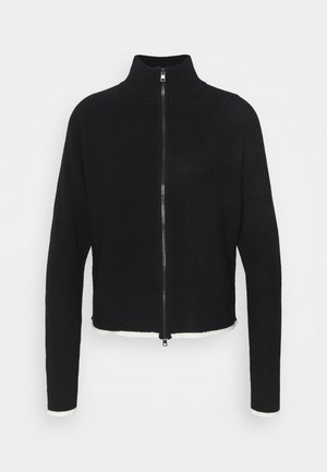 Schwarze, gerippte Strickjacke mit Reißverschluss, hohem Kragen und weißen Abschlüssen an Bündchen und Saum, vor schlichtem Hintergrund dargestellt.