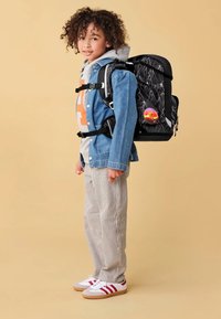 Child with curly hair wearing a denim jacket, striped pants, and sneakers, carrying a black backpack with a colorful round badge.