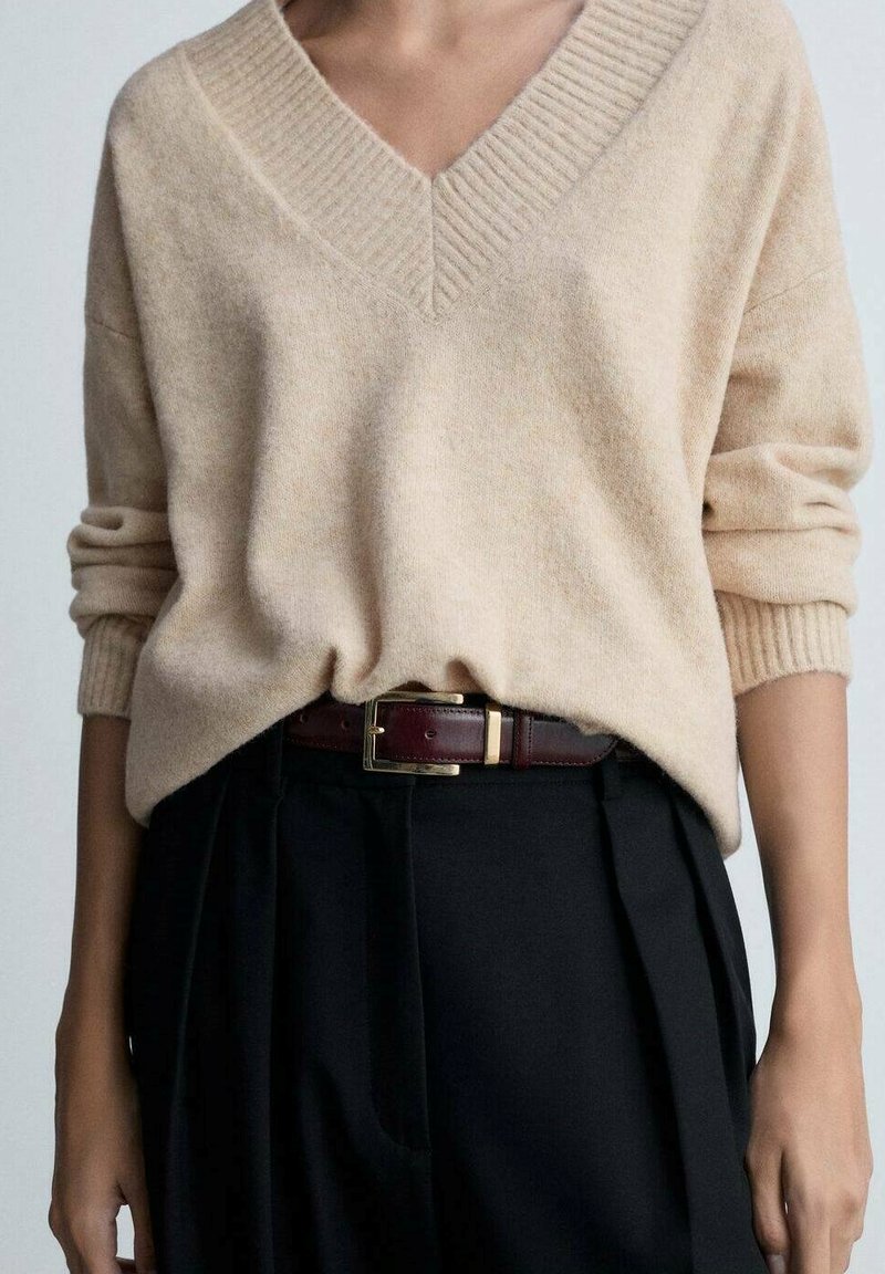 Maglione beige lavorato a maglia con scollatura a V profonda e bordi a costine, abbinato a pantaloni neri a pieghe e una cintura marrone scuro con fibbia dorata.