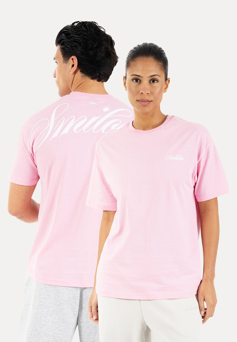 Homme et femme portant des t-shirts rose clair, femme de face, homme de dos montrant un grand texte blanc en cursive au dos.