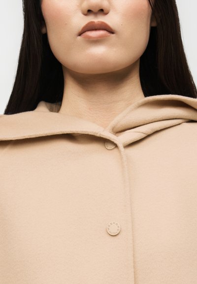 Γυναίκα με μακριά σκούρα μαλλιά που φοράει μπεζ μάλλινο παλτό με κουκούλα και στρογγυλά κουμπιά, με ορατό το κάτω μέρος του προσώπου και τον λαιμό της.