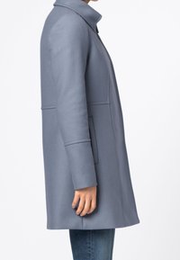 Cappotto grigio strutturato con collo alto, tessuto strutturato, tasche laterali e vestibilità sagomata, mostrato di profilo su uno sfondo uniforme.