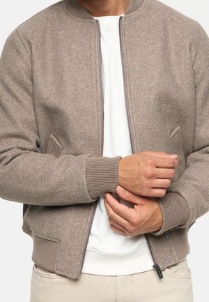 Uomo con giacca bomber in pile beige sopra una camicia bianca, che si sistema il polsino della giacca con entrambe le mani.
