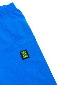 Niebieskie bawełniane szorty z elastycznym paskiem, z boczną kieszenią i czarną metką z napisem "ZONE 95 GAME" w zielonym kolorze.