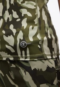 Κοντινό πλάνο αθλητικής ένδυσης με πράσινο και μπεζ καμουφλάζ, με εμφανείς ραφές και λογότυπο «C DRY» στο πάνω μέρος.