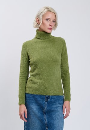 Kvinde med kort blondt hår bærer en grøn rullekravesweater og blå jeans, står mod en ensfarvet lys baggrund og kigger fremad.
