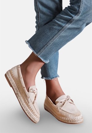 Pieds chaussés d'espadrilles beiges à enfiler avec des lacets, assorties à un jean bleu court aux ourlets effilochés.