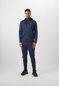 Marine atletisk treningsdrakt med blå detaljer, inkludert en hettejakke med glidelås og matchende bukser. Laget av elastisk, pustende stoff.