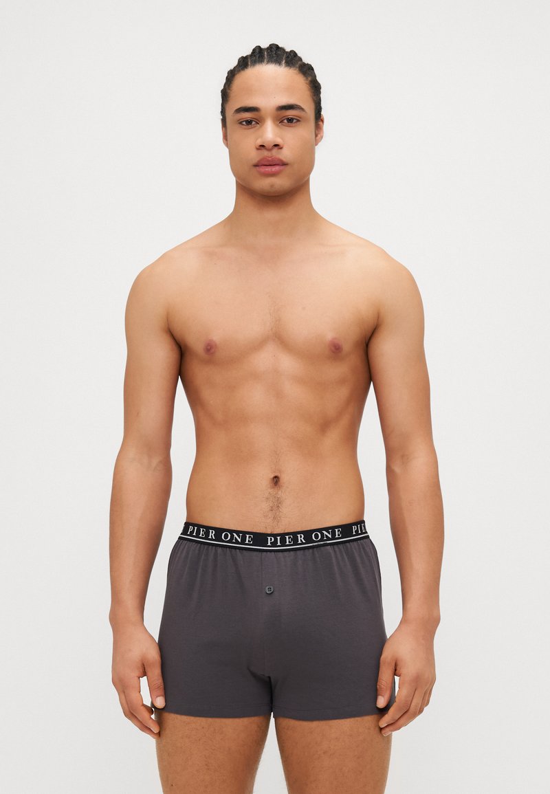 Graue Boxershorts mit schwarzem Bund, der den Text "PIER ONE" trägt. Glatte Textur und Knopfverschluss vorne. Das Modell zeigt den Oberkörper.
