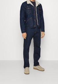 Džínová bunda s kožešinovým límcem, tmavě modré džíny a béžové semišové boty. Bunda má kapsy a detailní švy.