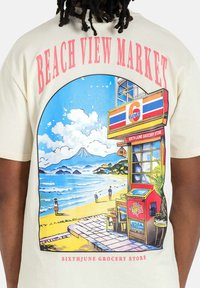 Crèmekleurig T-shirt met een grafische print van een strandscène, een gemakswinkel en bergen, met vetgedrukte tekst erboven.