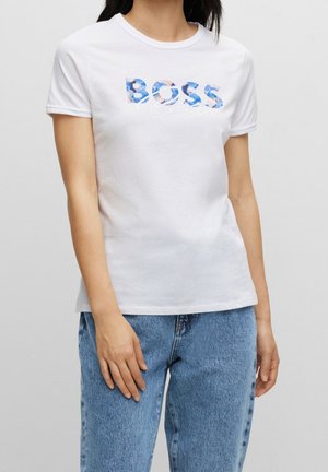 Kobieta ubrana w białą koszulkę z krótkim rękawem z kwiatowym nadrukiem "BOSS" oraz jasnoniebieskie dżinsy, stojąca na tle jednolitego szarego tła.