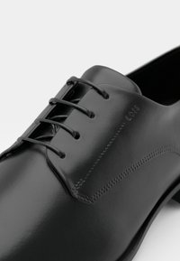 Chaussure habillée en cuir noir avec finition lisse, design à lacets et détails subtils de couture le long de la tige pour un raffinement supplémentaire.
