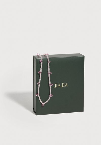 Collier délicat en perles avec petits pendentifs en forme de fleurs roses posé sur une boîte verte foncée de la marque JIAJIA sur fond blanc.