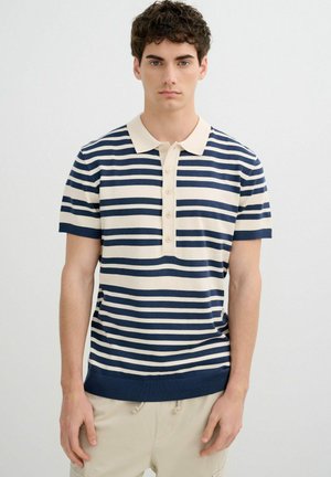 Joven de pie vistiendo una camiseta polo de manga corta con rayas horizontales en azul marino y crema y pantalones beige sobre un fondo liso.