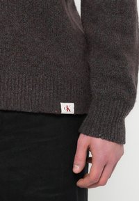 Sötétbarna kötött pulóver texturált mintával és színes pöttyökkel, bordázott mandzsettával és egy kis márkás címkével az alján.