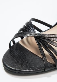 Sandale noire à brides avec motif texturé et plusieurs fines brides qui se croisent sur l'orteil ouvert, avec semelle intérieure beige visible.