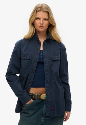 Navy blue button-up shirt in soft fabric with a collar, two front pockets, and brown buttons, worn over a dark blue top and denim jeans.